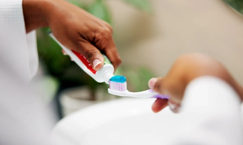 A close-up shot shows a person applying a blob of blue toothpaste onto a toothbrush in a bathroom.